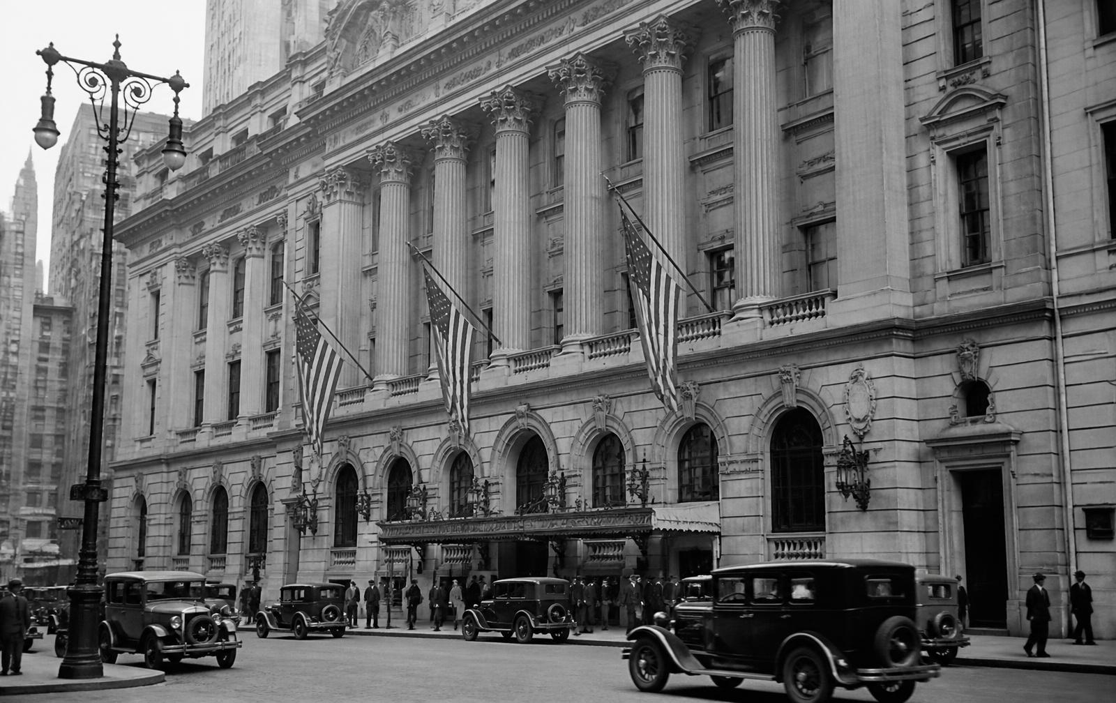 Archival view of a Beaux-Arts civic building in 1920s New York with flags and period automobiles